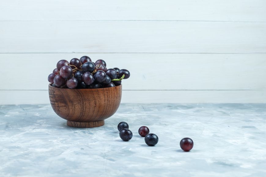 A wooden bowl filled with dark purple grapes sits on a light gray surface, with a few grapes scattered outside the bowl. The background is white wooden paneling.
