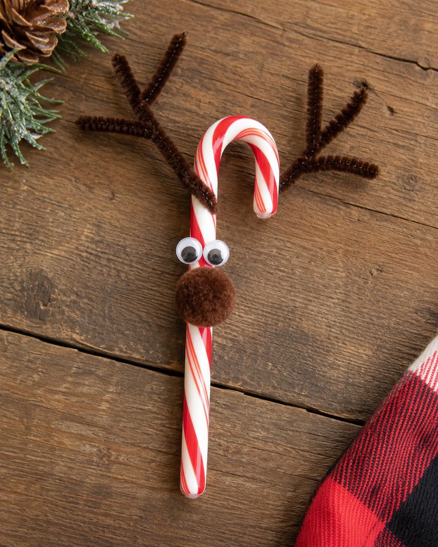 A candy cane decorated as a reindeer with googly eyes, a brown pom-pom nose, and pipe cleaner antlers, placed on a wooden surface next to greenery and a red plaid cloth.