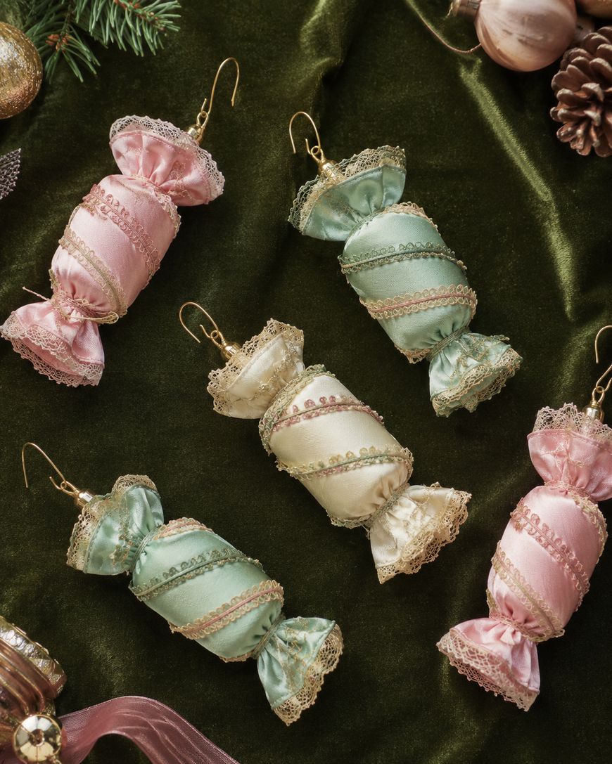 Five pastel-colored, candy-shaped ornaments with lace and ribbon details rest on a dark green velvet fabric, surrounded by pinecones and Christmas decorations. Each ornament has a gold hook for hanging.