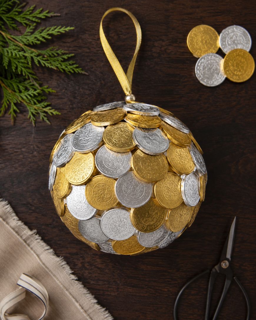 A round ornament covered with gold and silver chocolate coins sits on a dark wooden table, surrounded by a ribbon, fabric, scissors, and greenery.