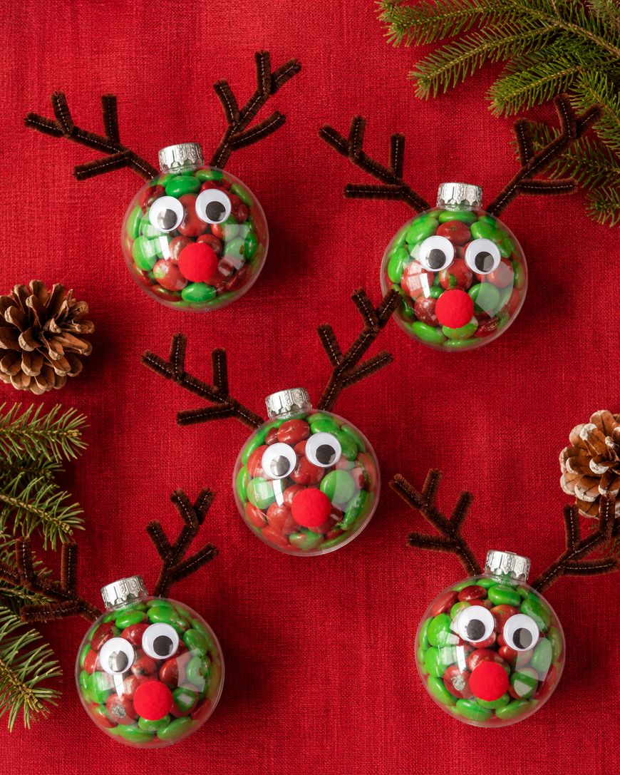 Five reindeer ornaments filled with red and green candies, decorated with googly eyes, red pom-pom noses, and brown pipe cleaner antlers, are arranged on a red cloth with pinecones and evergreen sprigs.