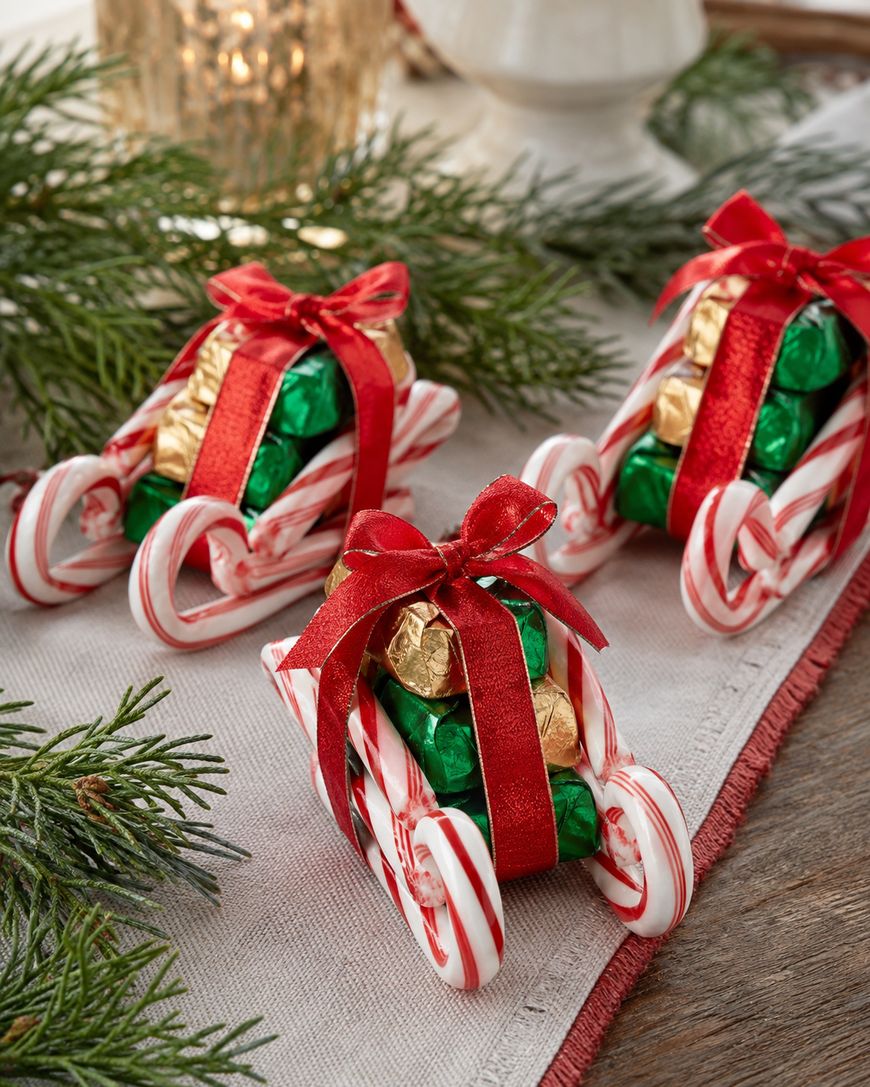 Three sleds made from candy canes, tied with red ribbons and topped with green and gold wrapped chocolates, are displayed on a festive table with pine branches and a candle in the background.