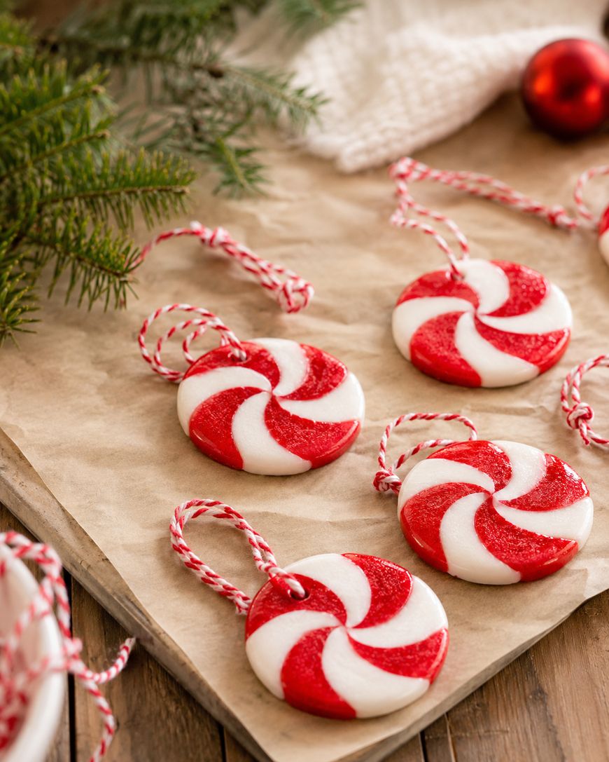 Red and white peppermint swirl ornaments with red and white string lie on brown paper, next to green pine branches and a white knitted cloth, creating a festive holiday scene.