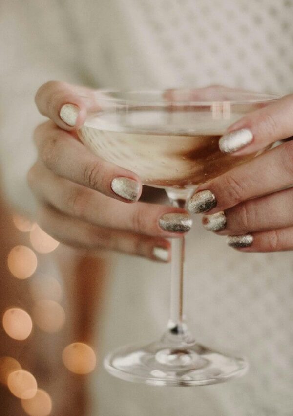 Close-up of hands with metallic silver nail polish holding a coupe glass filled with a light-colored drink. Warm, blurry lights and a textured sweater are visible in the background.