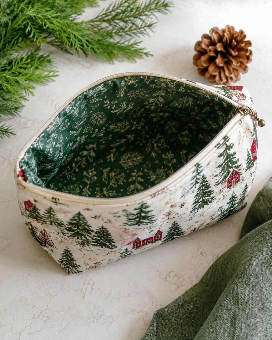 A zippered fabric pouch with Christmas-themed houses and trees sits open on a table, revealing a green, leafy-patterned lining inside. Pine branches, a pinecone, and a green cloth are nearby.