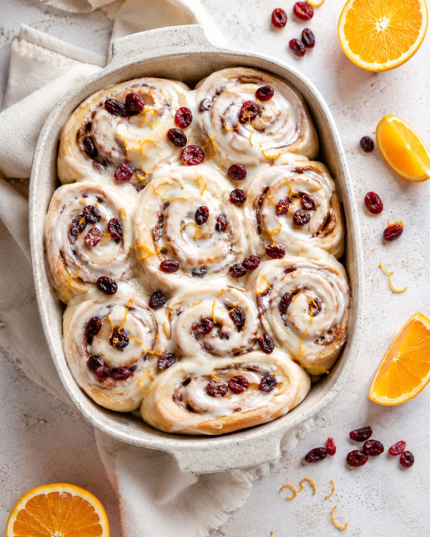 A baking dish filled with nine frosted cinnamon rolls, topped with dried cranberries and orange zest. Slices of fresh orange and scattered cranberries are nearby on a light surface.