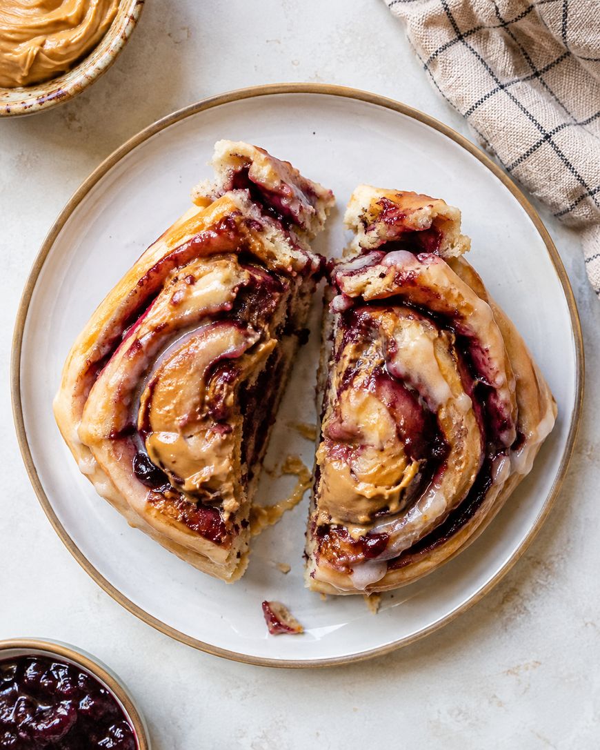 A large cinnamon roll topped with peanut butter and jelly is split in half on a white plate, with bowls of extra peanut butter and jelly nearby, and a checkered cloth in the corner.