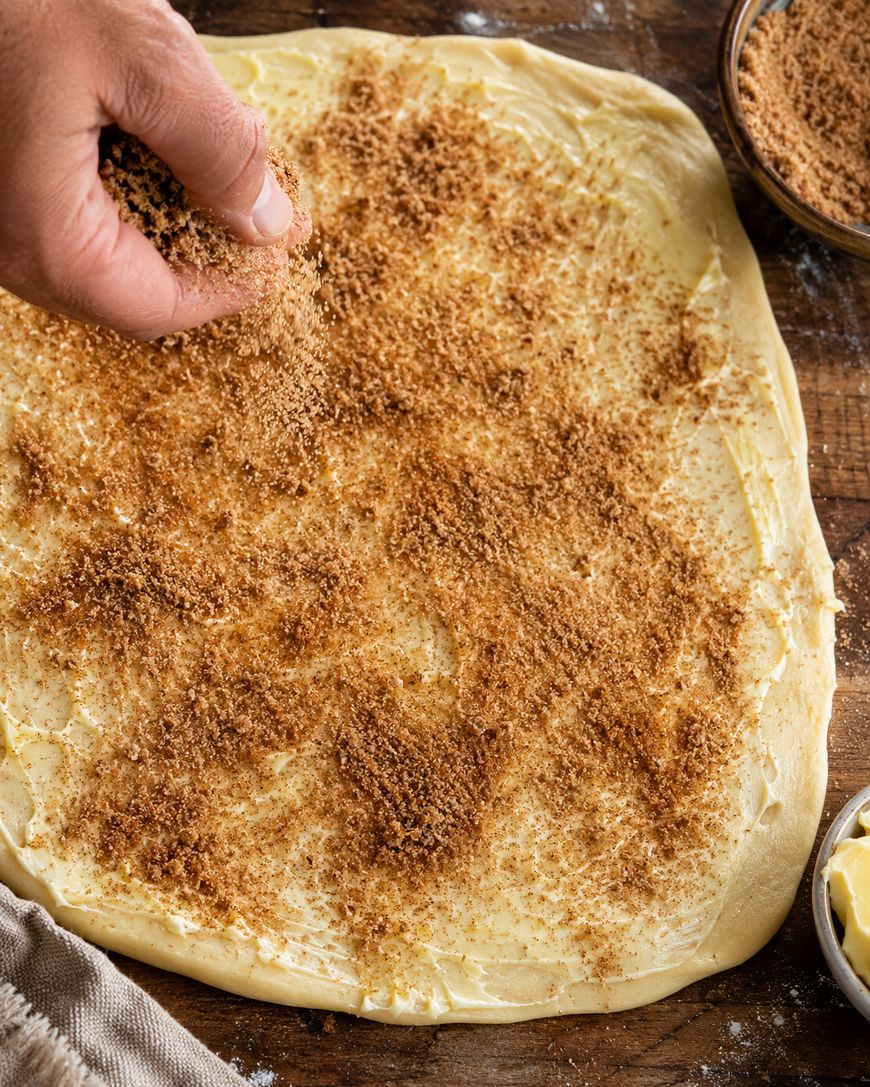 A hand sprinkles a brown sugar and cinnamon mixture over rolled-out dough on a wooden surface, preparing it for baking. Bowls with ingredients are nearby.