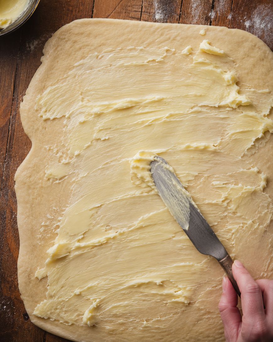 A hand spreads softened butter over a large, rolled-out rectangle of dough on a wooden surface, using a butter knife.