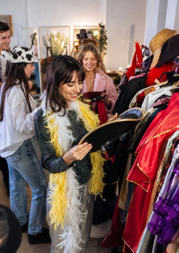 Four people are shopping in a vintage clothing store, smiling and browsing colorful costumes on racks. One woman in front wears a yellow feather boa and holds a hat, while others chat and pick out outfits.