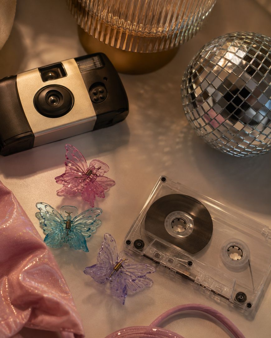 A retro camera, cassette tape, disco ball, pink scrunchie, and three butterfly hair clips in blue, pink, and purple lie on a shiny surface under warm light.