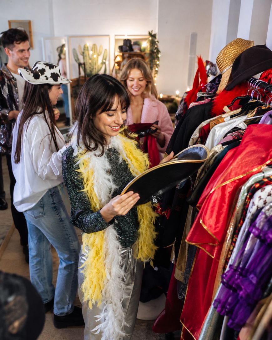 A smiling woman wearing a yellow feather boa looks at a hat in a vintage clothing store, surrounded by racks of colorful clothes and three other people browsing.