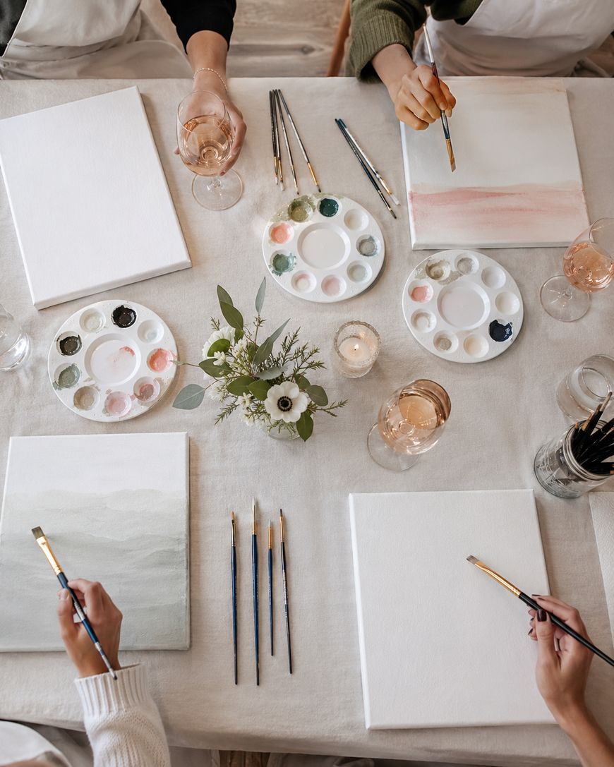 Four people sit around a table with blank canvases, paint palettes, brushes, and glasses of wine, painting soft abstract landscapes. A small floral centerpiece and candles decorate the table.