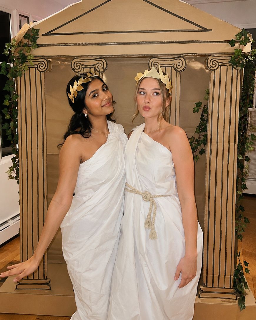 Two women wear white togas and gold leaf headbands, posing in front of a cardboard backdrop painted to look like a Greek temple with columns and ivy decorations.