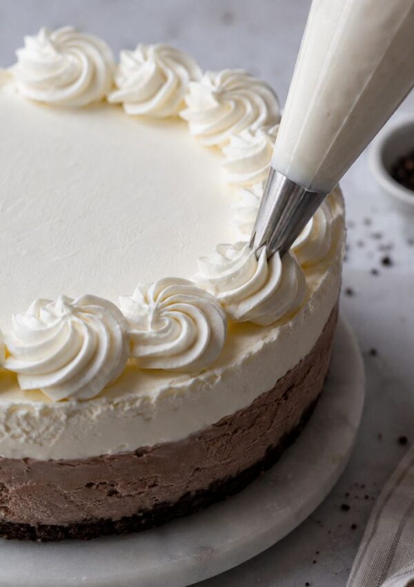 A hand pipes swirls of whipped cream onto the edge of a layered chocolate and vanilla cheesecake on a marble cake stand, with a bowl and napkin in the background.