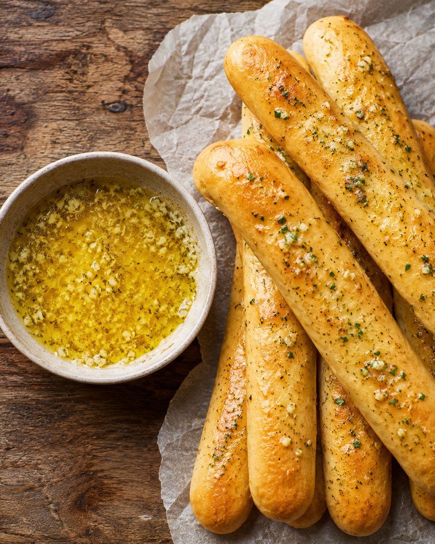 A pile of golden, seasoned breadsticks on parchment paper next to a bowl of garlic butter dipping sauce, set on a rustic wooden surface.
