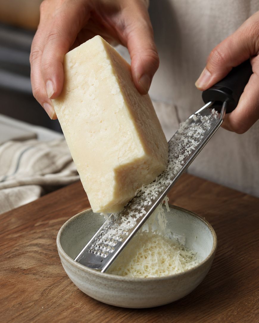 A hand grates a block of hard cheese, such as Parmesan, over a small bowl, collecting fine cheese shreds on a wooden surface.