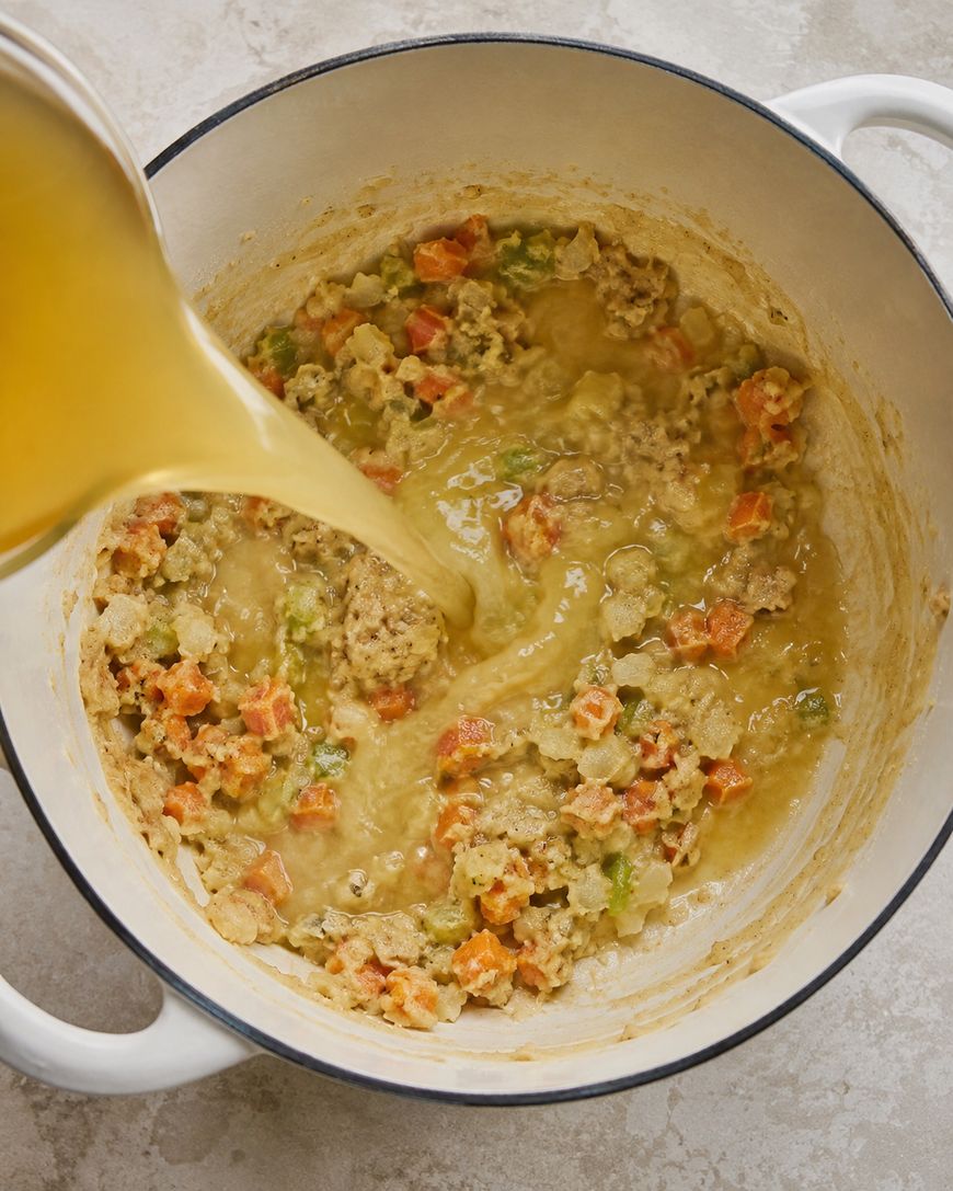 Broth is being poured into a white pot containing a mixture of sautéed carrots, celery, onions, and cooked ground meat on a light-colored countertop.