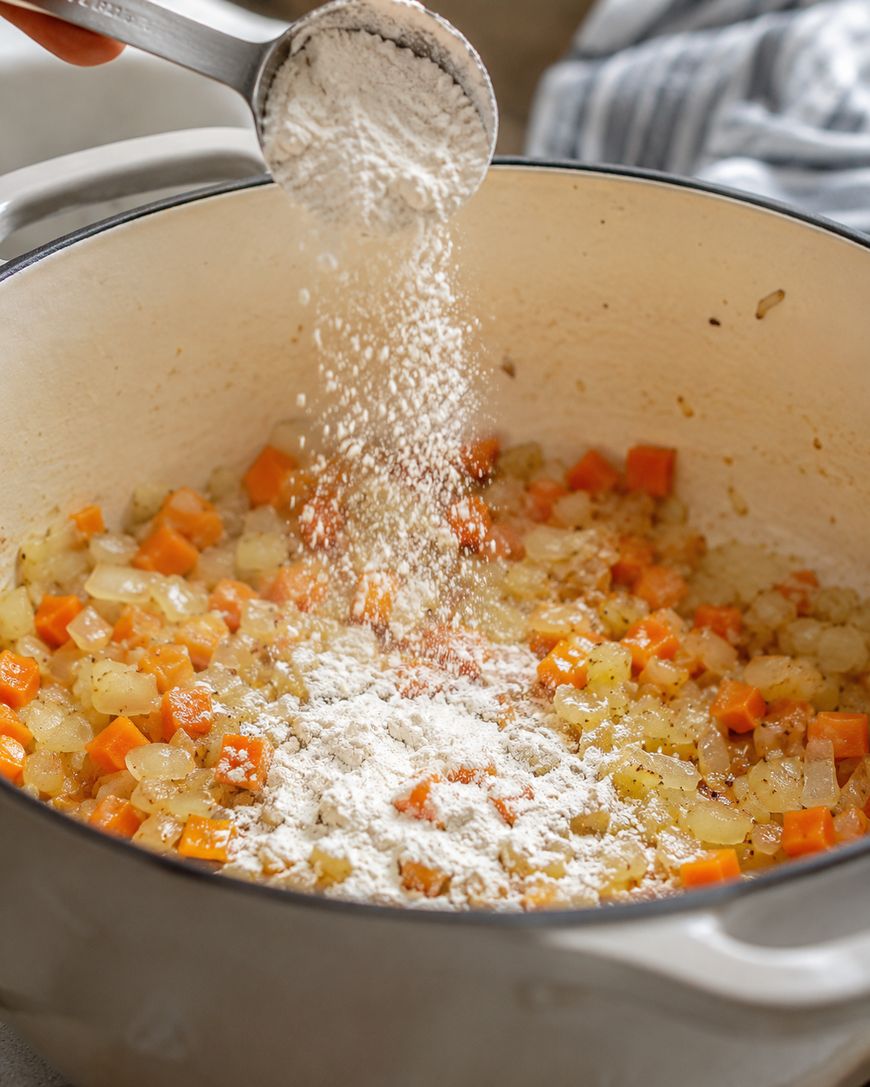 A hand sprinkles flour from a measuring cup into a pot of sautéed onions and carrots, preparing the base for a recipe.