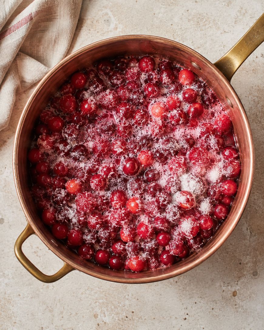 A copper saucepan filled with cranberries and sugar sits on a light countertop, with a beige towel partially visible in the upper left corner.