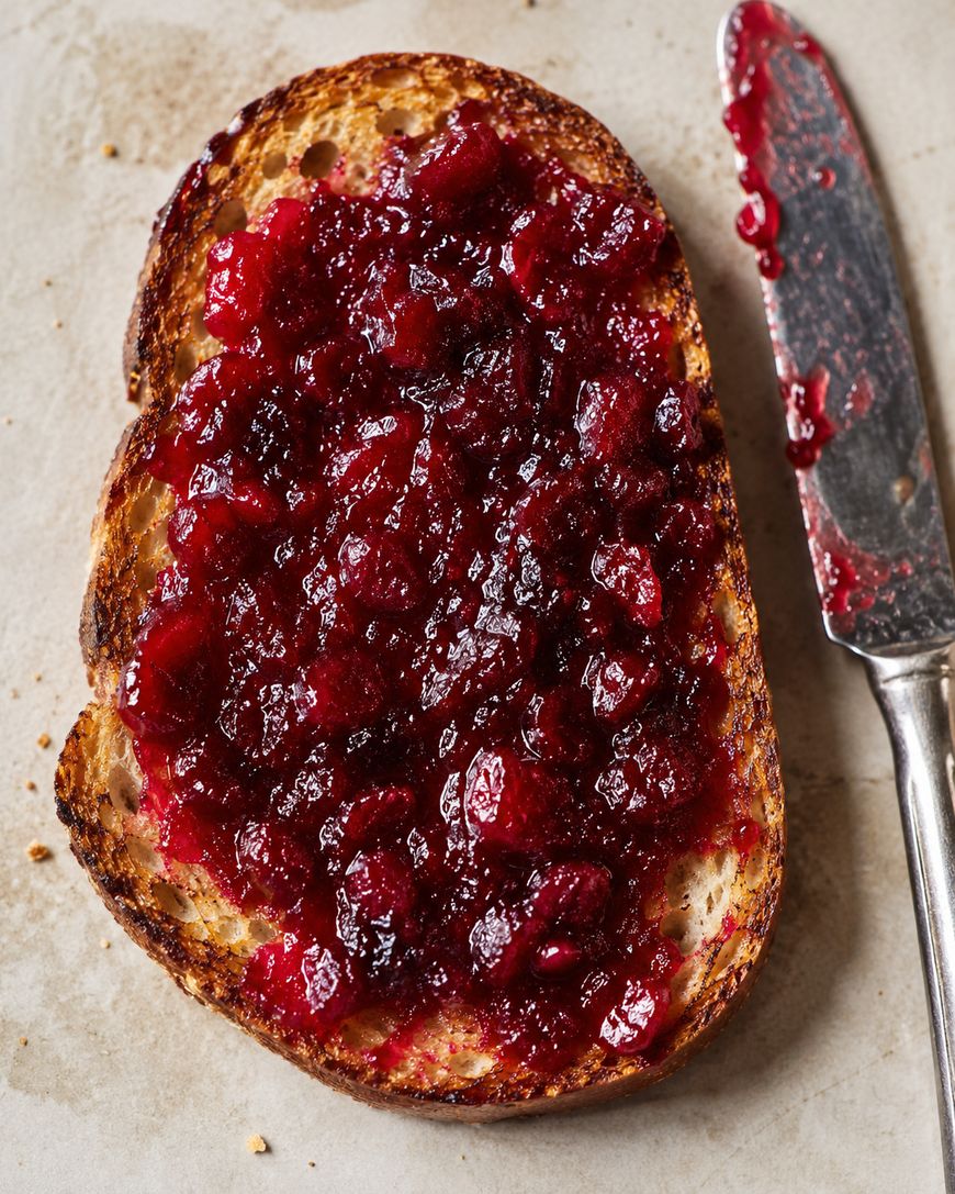 A slice of toasted bread topped with chunky red jam sits on a light surface next to a butter knife with jam residue on its blade.