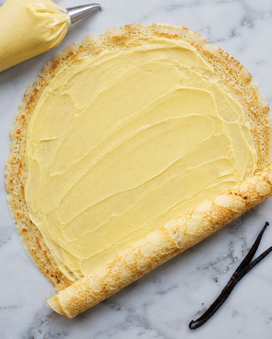 A thin crepe is being rolled up with a layer of creamy filling. A piping bag with more filling and a split vanilla bean pod are nearby on a marble surface.