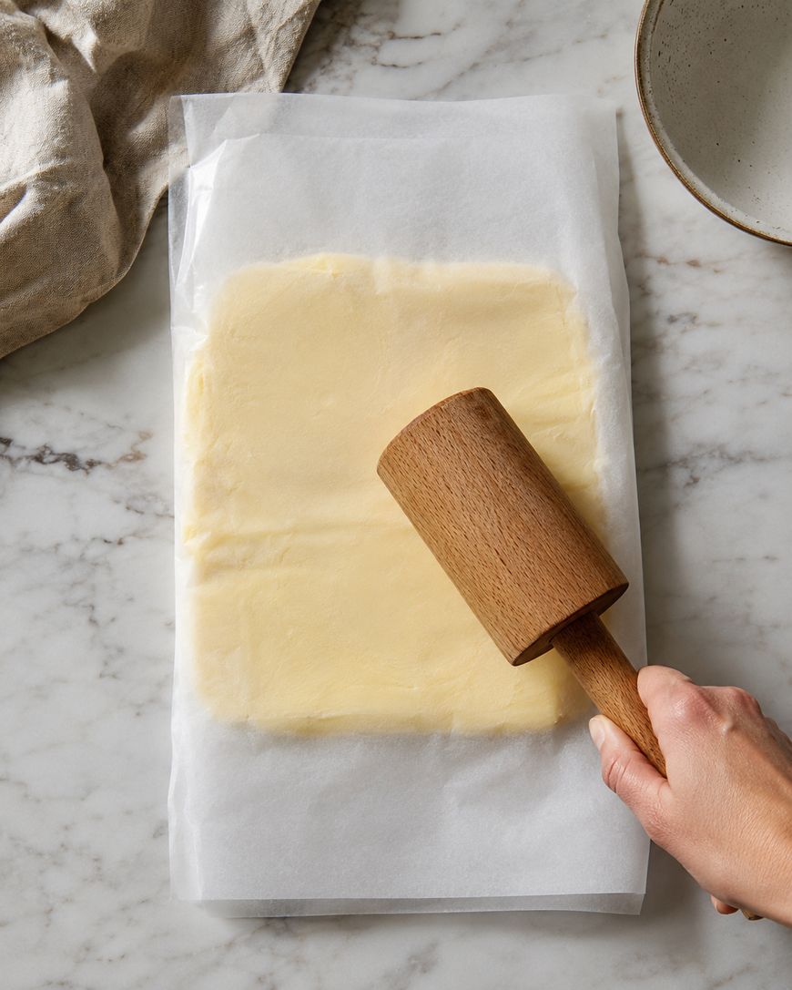 A hand uses a wooden rolling pin to flatten butter between sheets of parchment paper on a marble countertop.
