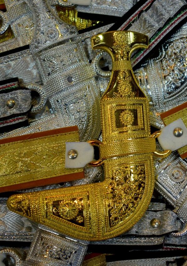 A golden, intricately decorated traditional curved dagger, known as a jambiya, displayed among other silver-colored daggers and ornate belts.