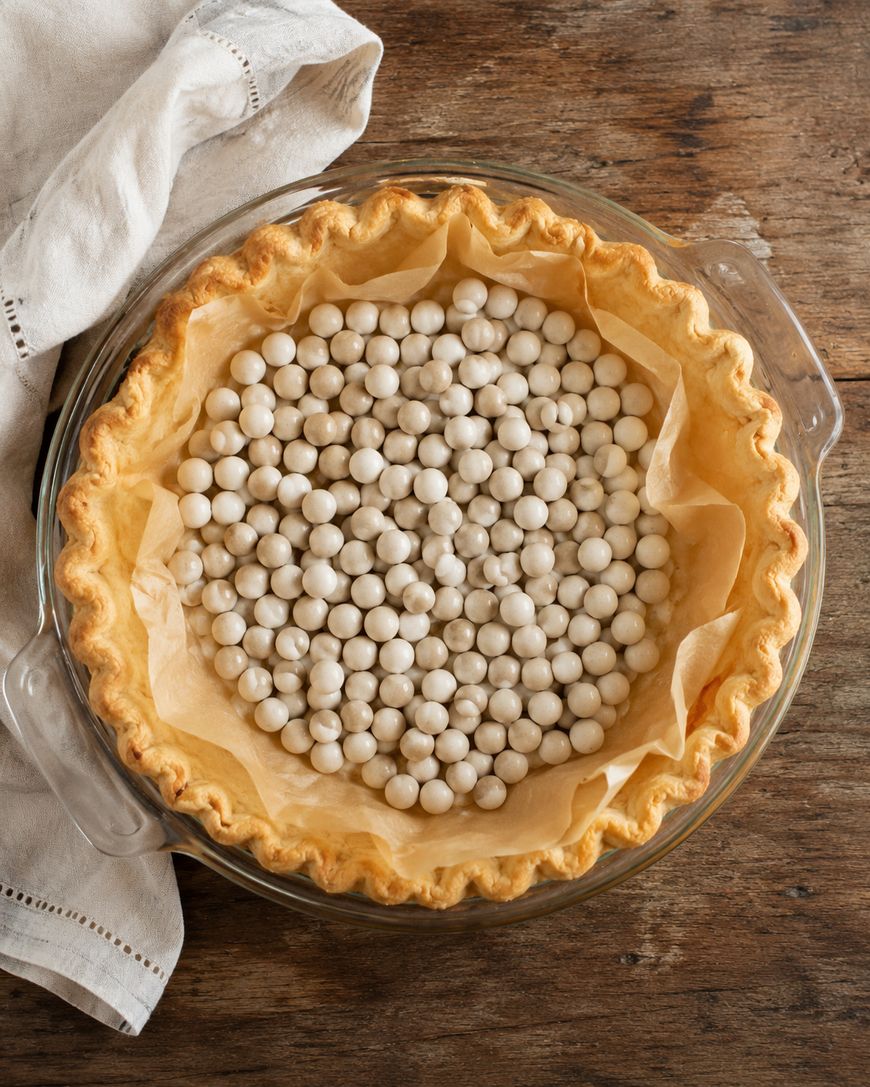 A pie crust in a glass dish lined with parchment paper and filled with white ceramic pie weights, sitting on a rustic wooden surface next to a folded beige cloth.