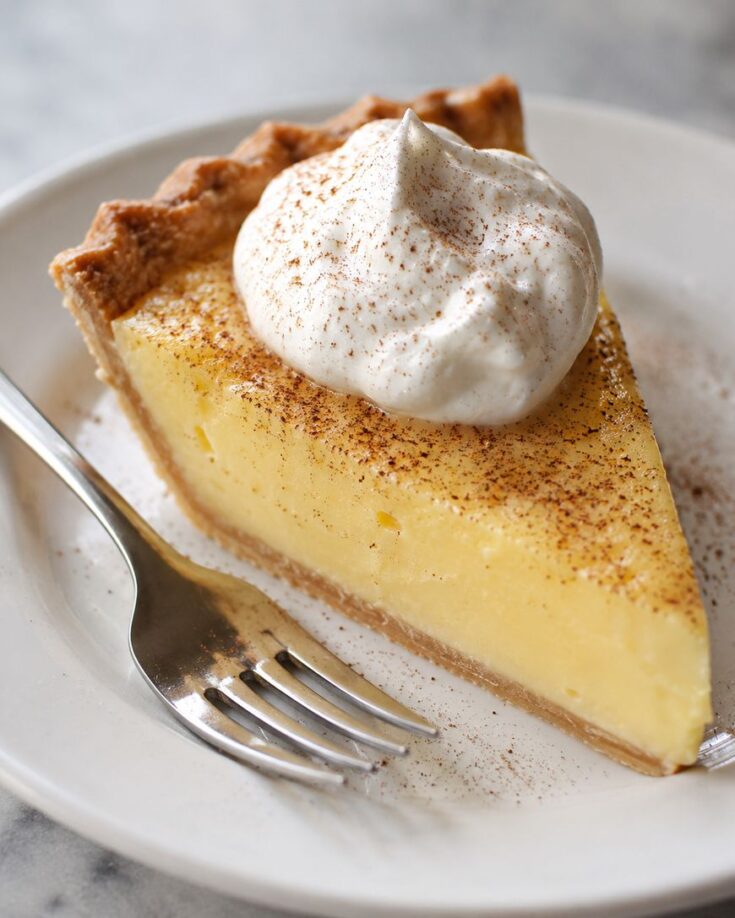 A slice of creamy custard pie with a golden crust, topped with a dollop of whipped cream and a sprinkling of cinnamon, sits on a white plate next to a fork.