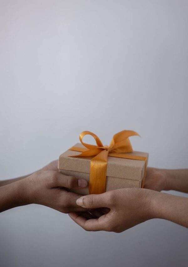 Two hands are giving a small, brown gift box with an orange ribbon to another pair of hands against a plain, light background.