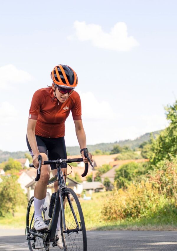 A cyclist wearing an orange helmet and jersey rides a bike uphill on a sunny day, with houses and greenery visible in the background.