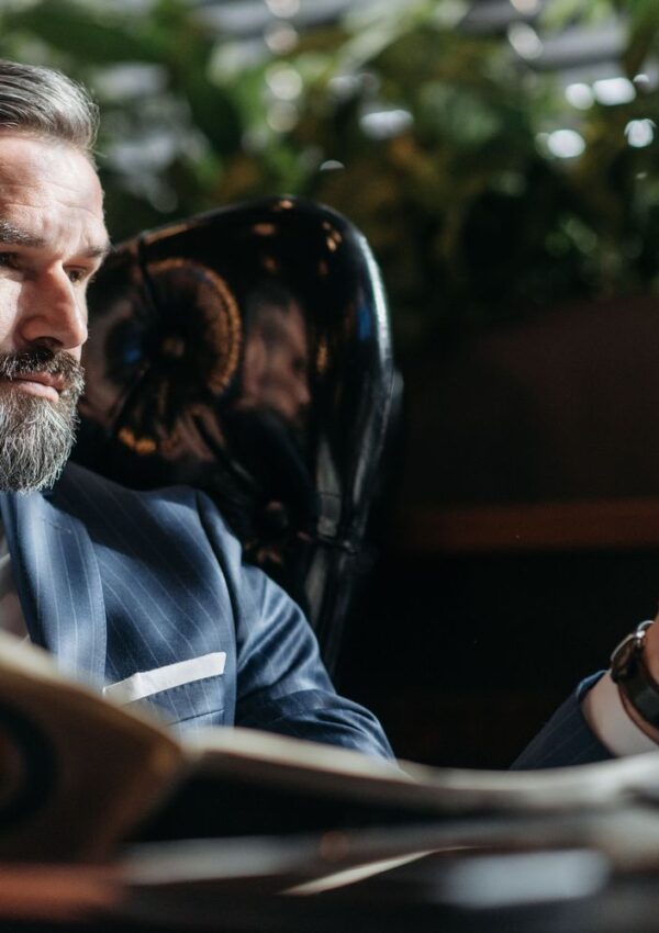 A man in a pinstripe suit with a beard sits in a chair, holding a glass and looking at it thoughtfully—a perfect reflection of personal style. Green plants appear blurred in the background, adding an elegant touch to this sophisticated date night wardrobe inspiration.