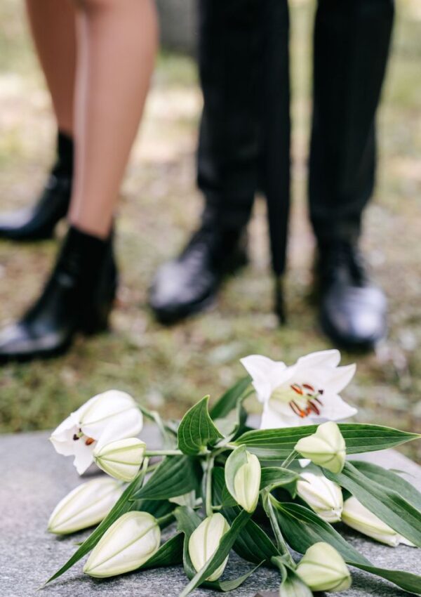 White lilies rest on a gravestone with two people standing nearby, only their legs and feet visible, suggesting a somber, mourning atmosphere at a gravesite.