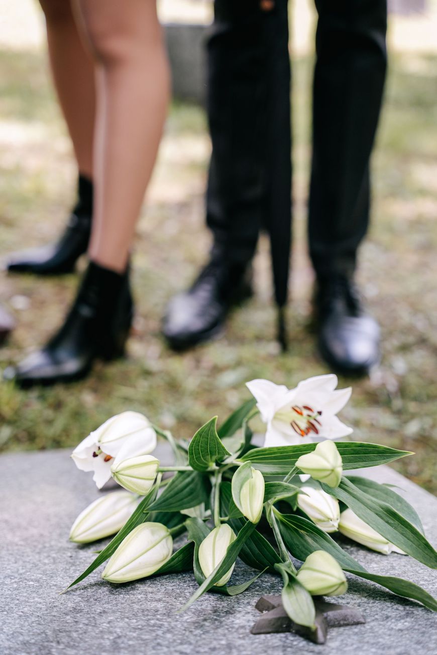 Two people in black stand by a grave covered with white lilies and greenery. Only their legs and shoes are visible, suggesting a somber funeral or memorial sceneāan emotional moment when thoughts might turn to important tasks ahead or reviewing a death notification checklist.