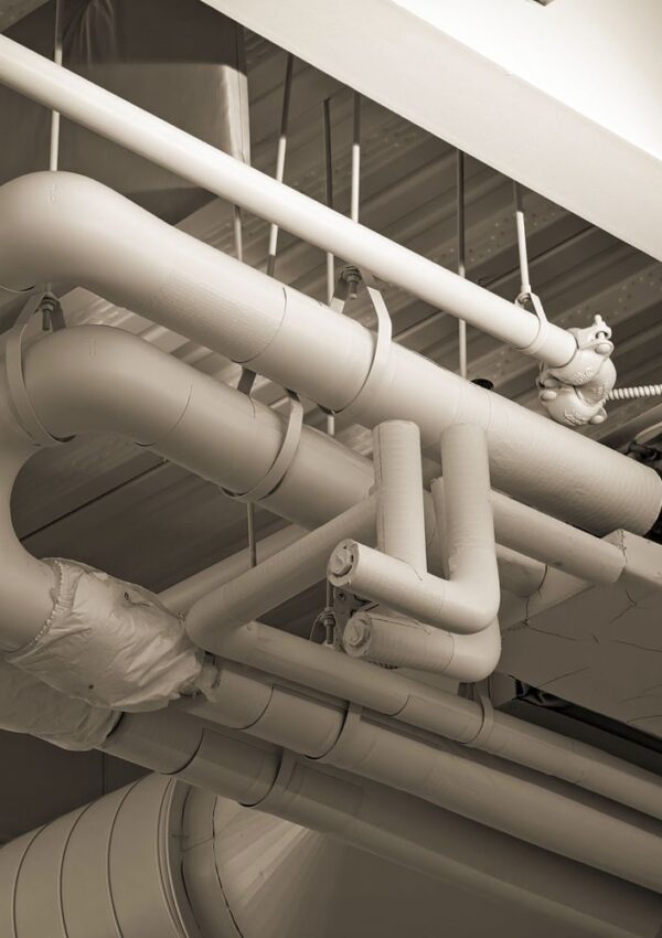 A network of large, insulated industrial pipes runs along a ceiling, with joints, bends, and valves visible—reminiscent of the systems found in historic Charleston homes. The image has a sepia tone, subtly highlighting the intricate details that can often conceal signs of dirty air ducts.