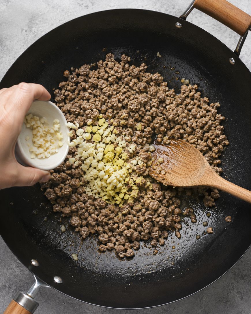 Ground beef is cooking in a black skillet, being stirred with a wooden spoon. A hand is adding chopped garlic and ginger from a small white bowl onto the beef.