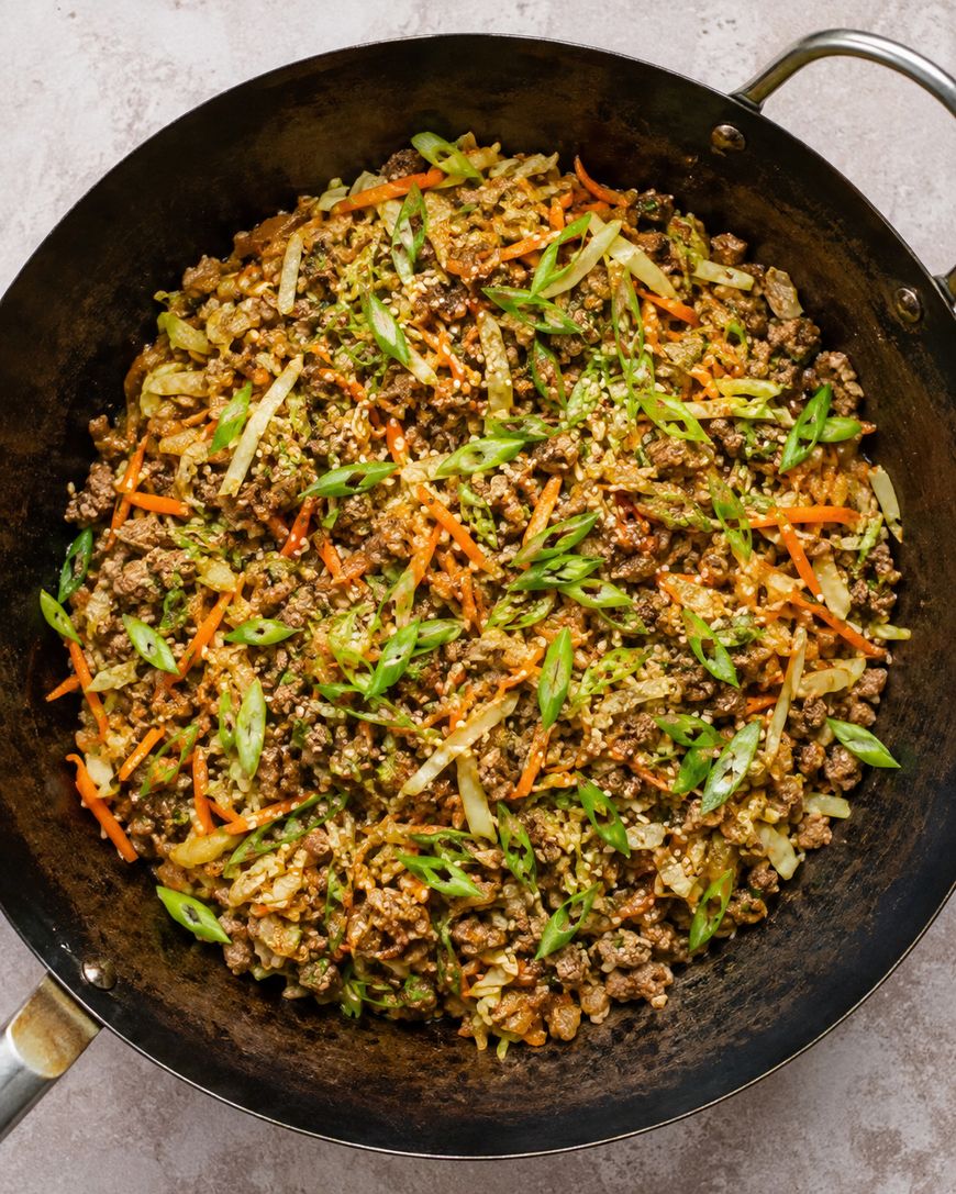 Ground meat, shredded carrots, cabbage, and green onions are stir-fried in a large black wok, creating a colorful and textured dish with a mix of vegetables and savory meat.