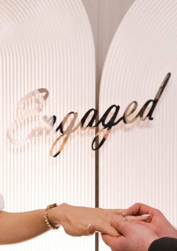 A person places a ring on anothers finger, with the word Engaged in elegant script in the background.