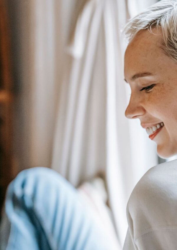 A person with short blonde hair and a white shirt smiles while sitting indoors, with soft natural light and neutral curtains in the background.
