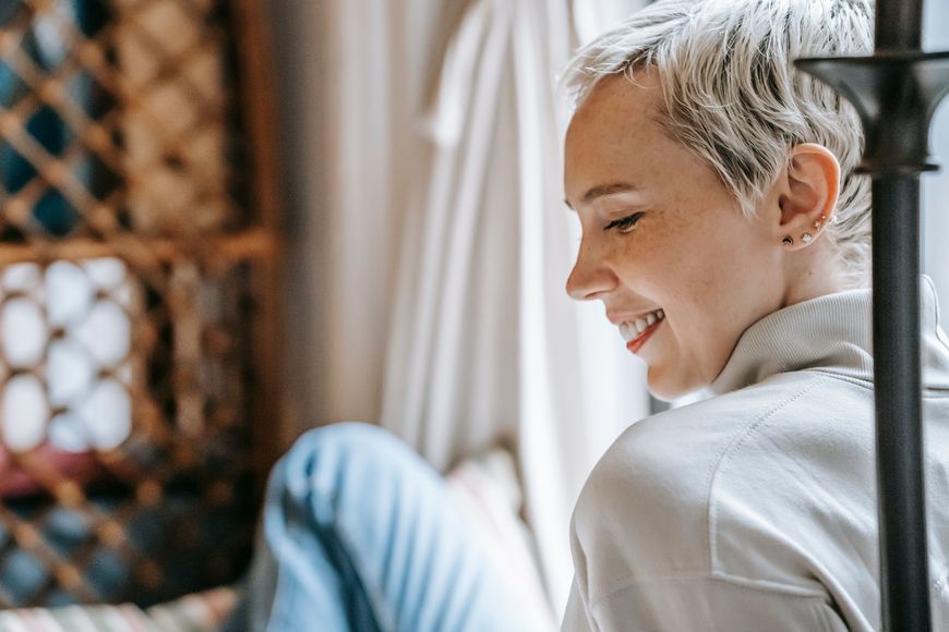 A person with short, light hair and a light sweater smiles while sitting indoors near a window, with soft natural light illuminating their face.