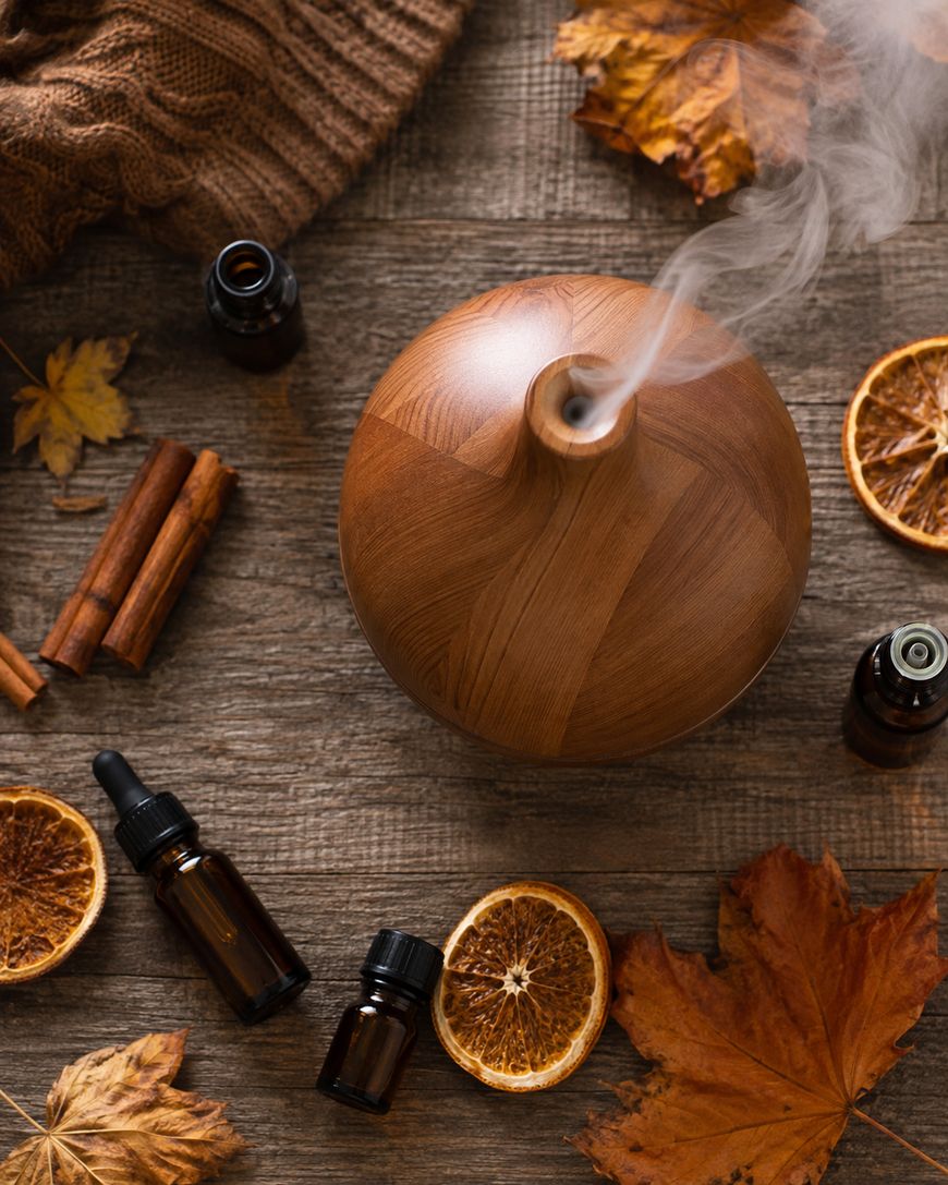 A wooden essential oil diffuser emits steam on a rustic table, surrounded by amber dropper bottles, dried orange slices, cinnamon sticks, autumn leaves, and a cozy knit fabric.