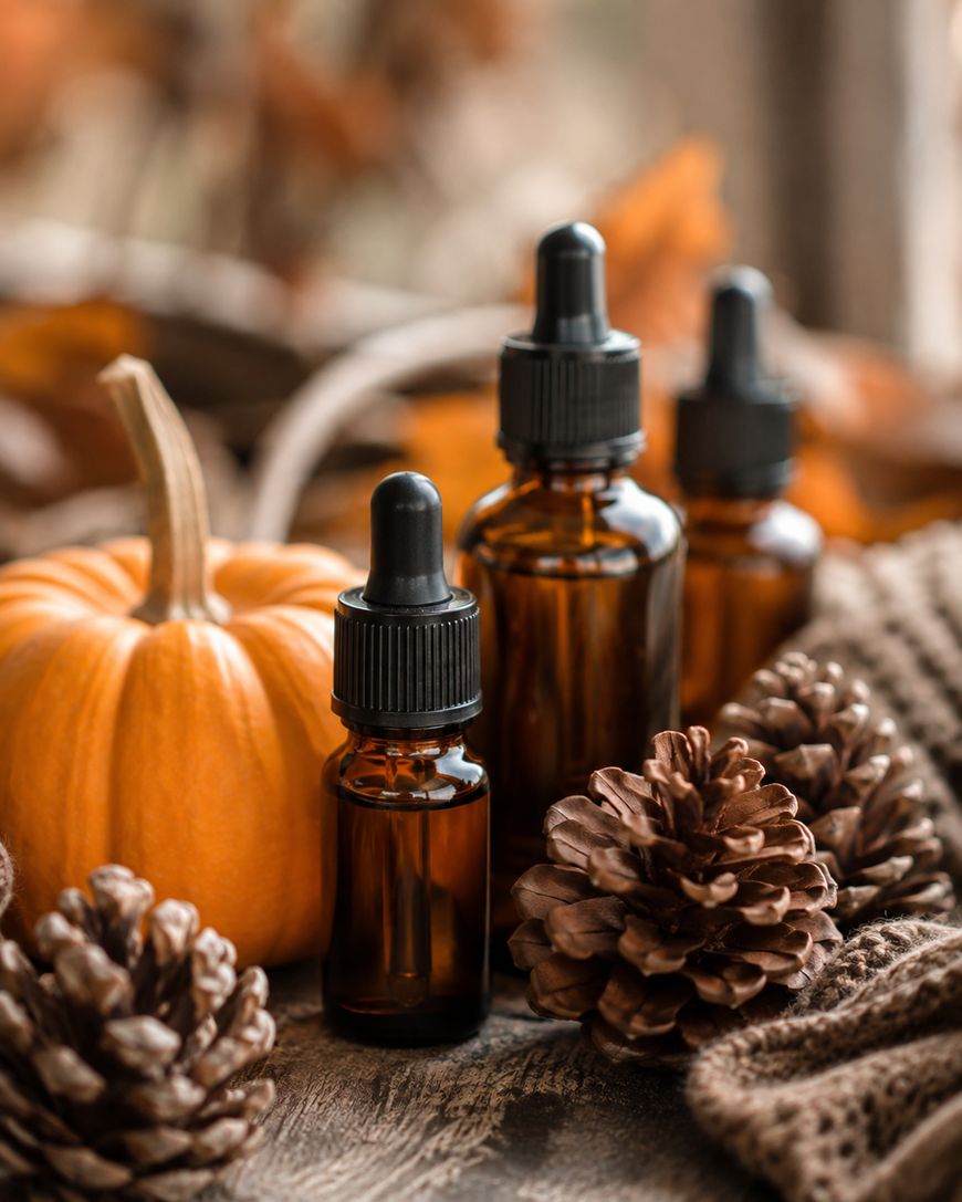 Three amber dropper bottles are arranged with pinecones, a small pumpkin, and a brown knitted fabric, creating an autumn-themed scene with warm colors and a rustic wooden surface.