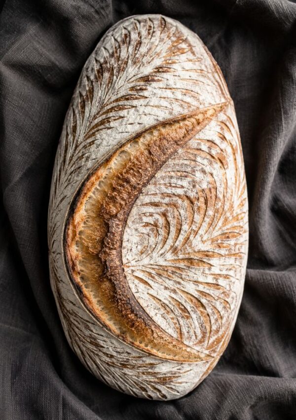 An oval sourdough loaf with intricate leaf patterns scored on the crust, resting on a dark cloth background.