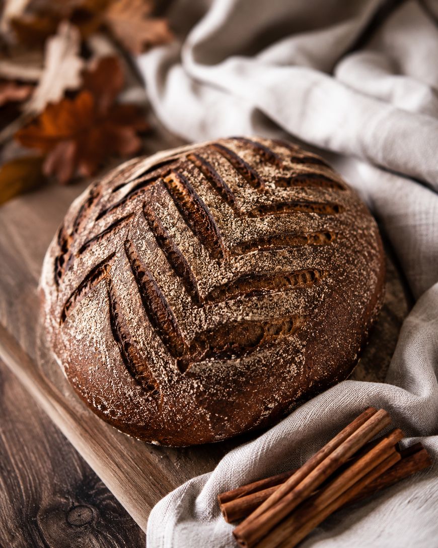 Round loaf of rustic bread with decorative scoring on top, resting on a wooden board next to a light cloth and cinnamon sticks; autumn leaves are in the background.