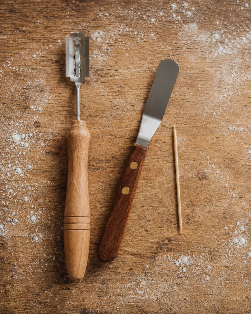 A bread lame, a butter knife with a wooden handle, and a toothpick are arranged on a floured wooden surface.