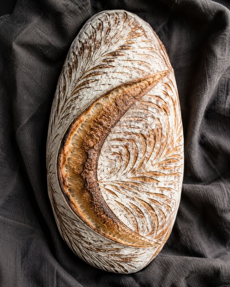 Oval loaf of artisan bread with intricate leaf and swirl patterns scored into the crust, resting on a dark, crumpled cloth.