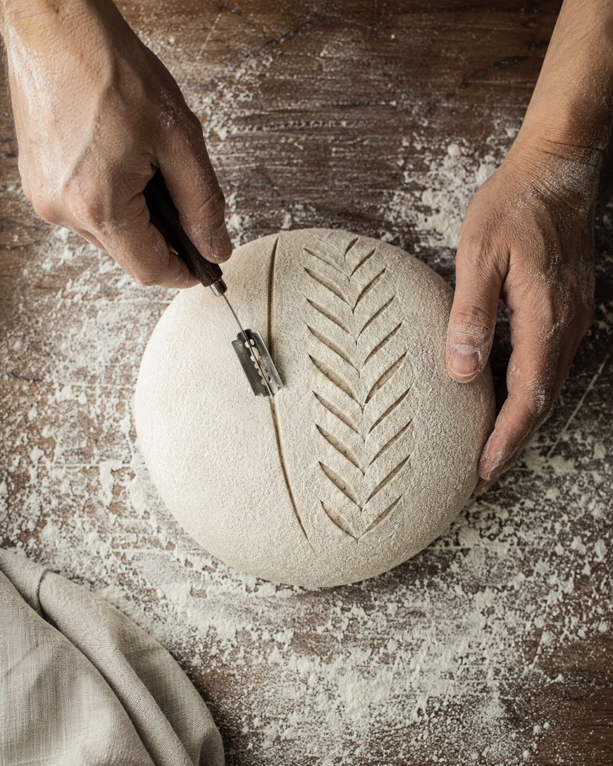 A person uses a razor blade to score patterns into a round loaf of dough on a floured wooden surface, preparing it for baking.