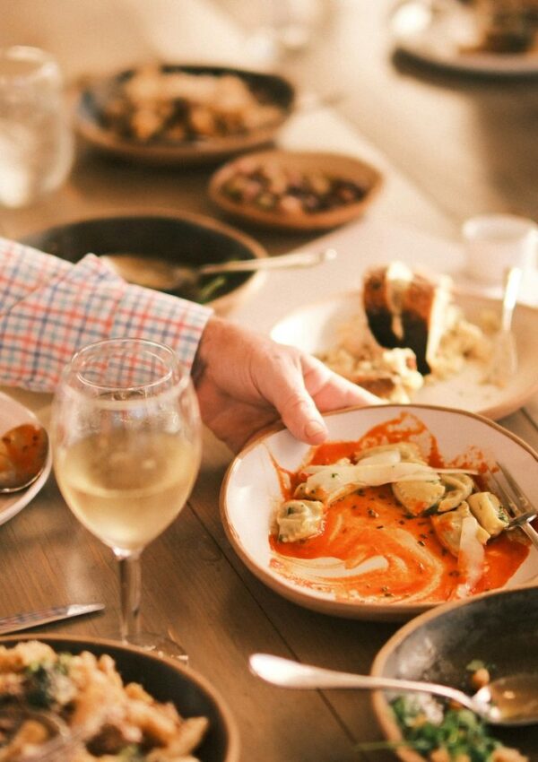 A hand holds a bowl of pasta in red sauce on a wooden table set with various dishes, including bread, salad, and a glass of white wine, highlighting the inviting presentation that elevates everyday meals.