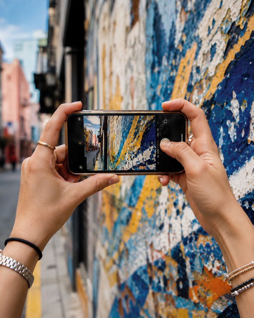 A person holds a smartphone to take a photo of a colorful, abstract mural on a city street wall, capturing the artwork closely. The persons hands and accessories are visible in the foreground.